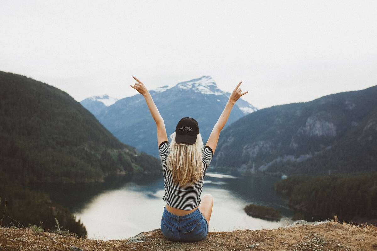 A woman with her back to the camera with her arms raised sitting in front of a mountain.